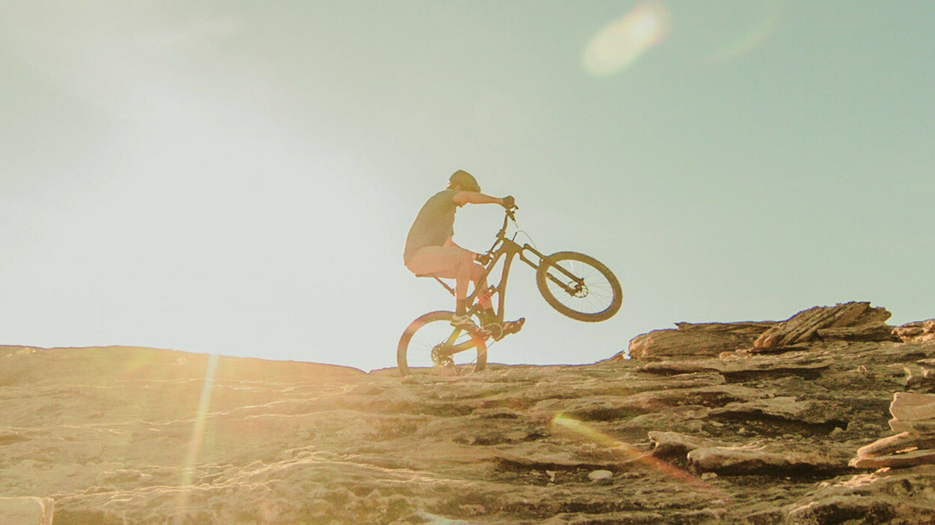 Mountainbiker fährt einen felsigen Anstieg hinauf bei Sonnenuntergang, mit sichtbaren Sonnenstrahlen im Hintergrund.