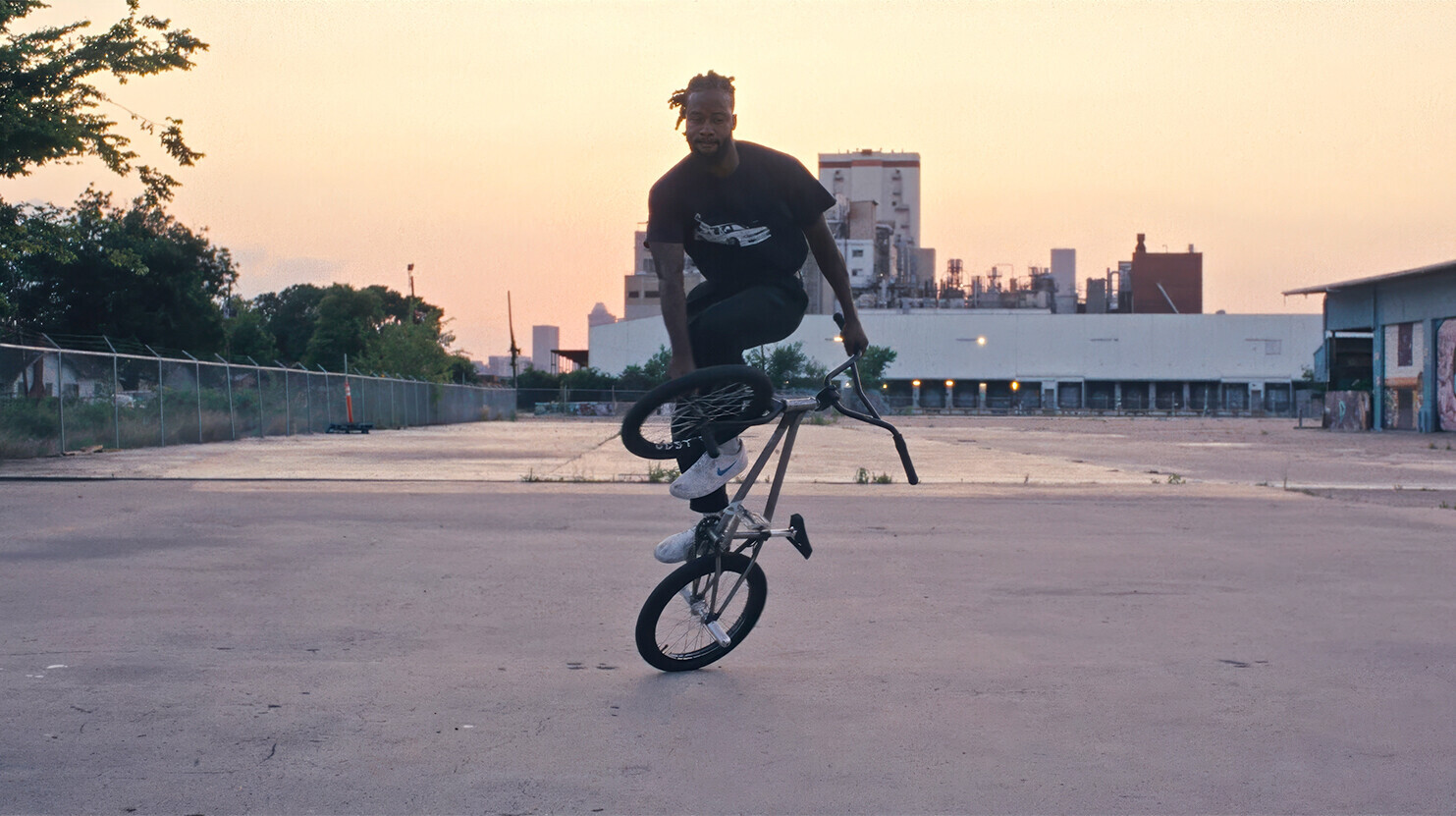 Omari Cato performing a flatland BMX trick in an urban industrial setting at sunset.