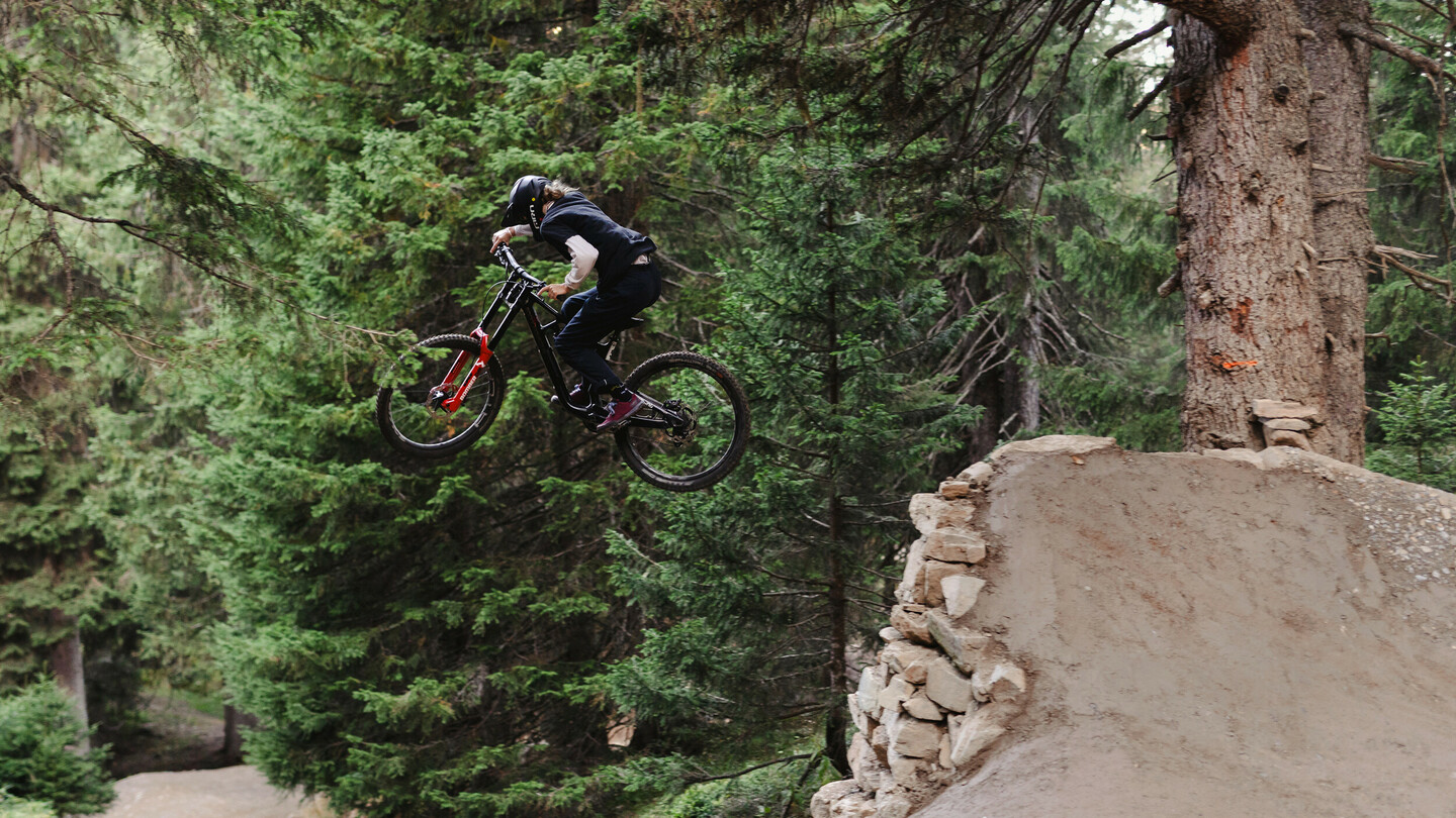 Ein Mountainbiker springt mit seinem Fahrrad von einer Rampe in einem dichten Wald. Der Fahrer trägt einen Helm und dunkle Kleidung, während er über die massive Erdhügelrampe fliegt.