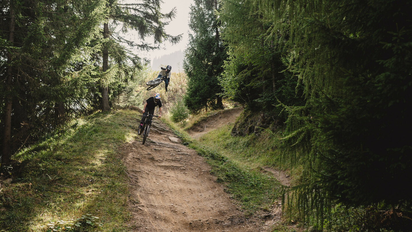 A mountain biker performs a high aerial trick while another rider navigates a forest trail. The scene is surrounded by tall trees and lush vegetation.