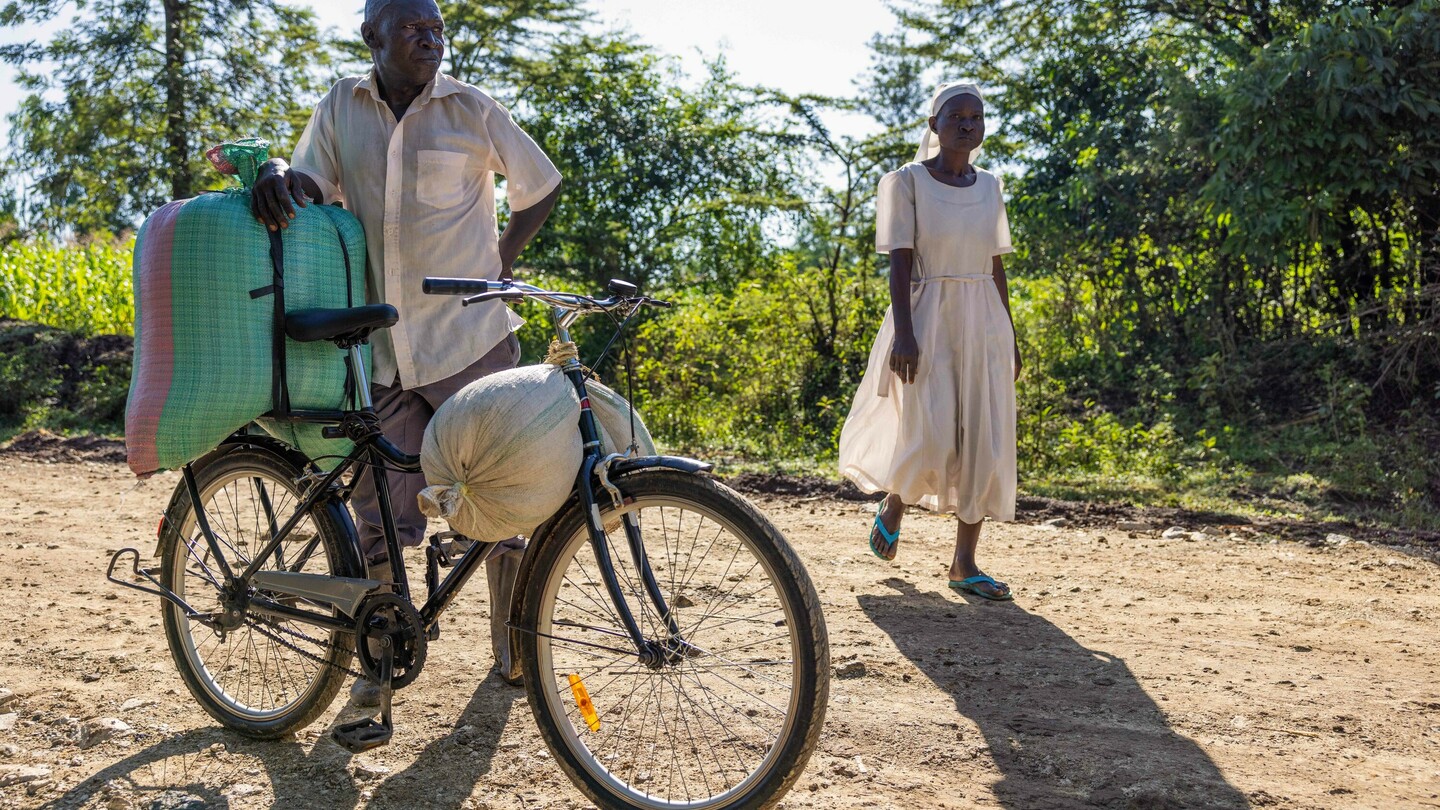 A man stands next to a loaded bicycle on a rural path, while a woman in a light dress walks by. Lush greenery is visible in the background.