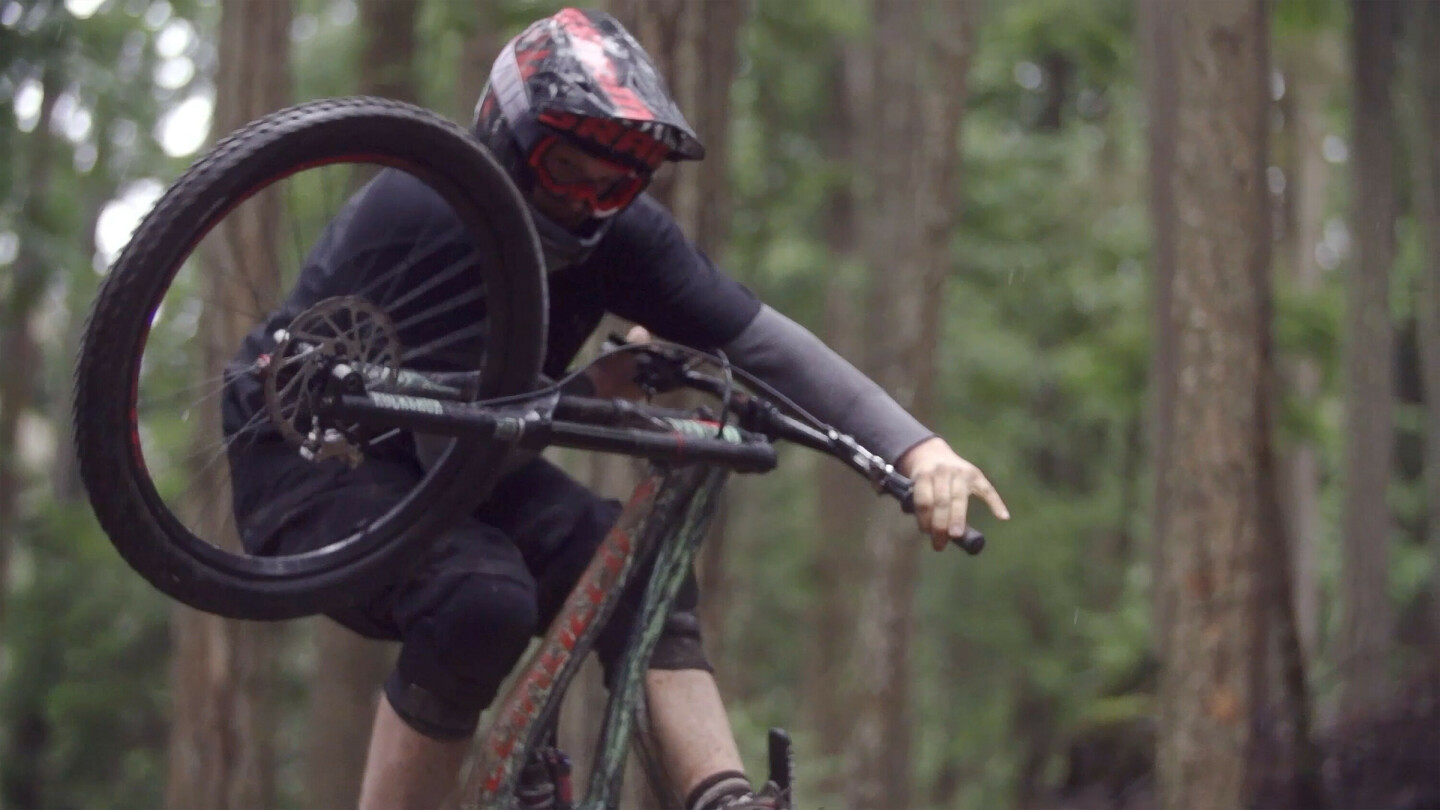 Mountain biker wearing a helmet and goggles performing a jump or trick in a forest, with the front wheel lifted.