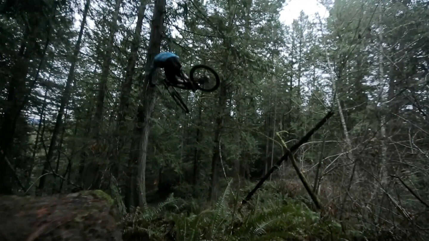 Ein Mountainbiker in der Luft während eines Sprungs in einem dichten Waldgebiet. Der Fahrer trägt dunkle Kleidung und der Wald ist voller hoher Bäume und dichtem Unterholz.