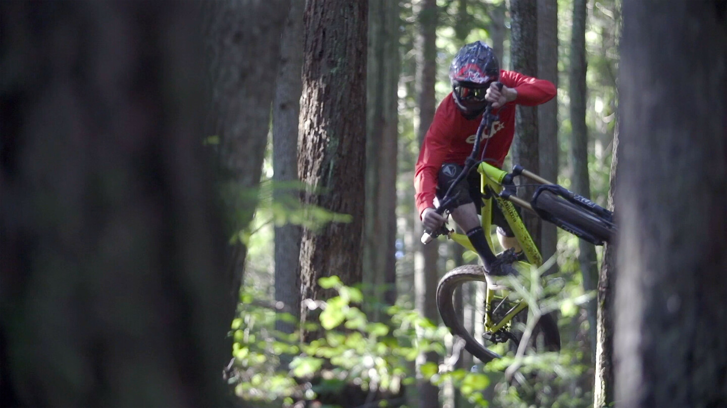 Mountainbiker mit Helm und rotem Shirt macht einen Sprung in einem dichten Wald, das Fahrrad ist dabei zur Seite geneigt.