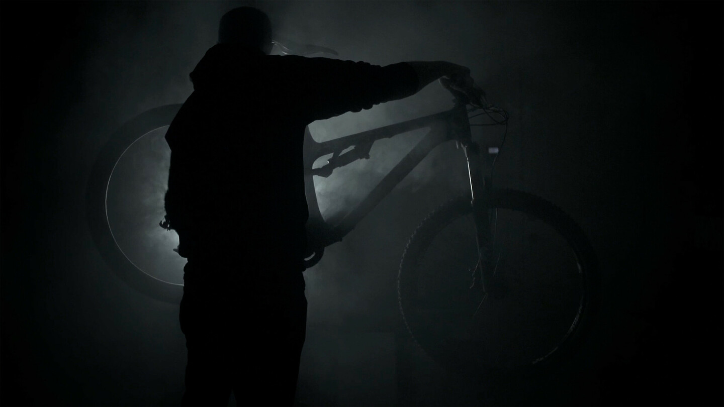 Silhouette einer Person, die ein Mountainbike in einer dunklen, rauchigen Umgebung hält, mit Licht von hinten.